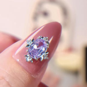 May include: A close-up of a silver ring featuring a large oval-shaped purple gemstone surrounded by smaller clear gemstones. The ring is on a finger with pink nail polish.