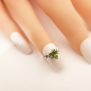 May include: A close-up of a fingernail with a green heart-shaped gem with silver wings attached to it.