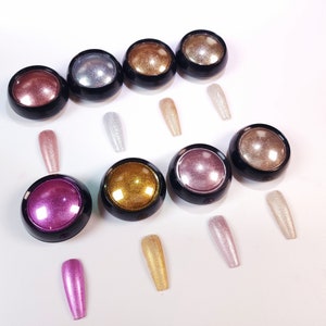 Puede incluir: Surtido de diez pequeños frascos redondos llenos de purpurina iridiscente en varios colores, incluyendo oro, plata, rosa y morado. Los frascos están dispuestos en dos filas sobre una superficie blanca.