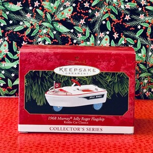 May include: A red and white box featuring a Hallmark Keepsake Ornament logo. The ornament is a white 1968 Murray Jolly Roger Flagship Kiddie Car Classic with red accents and blue wheels. The box reads "1968 Murray® Jolly Roger Flagship Kiddie Car Classics COLLECTOR'S SERIES".