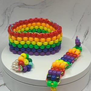 May include: A collection of colorful beaded accessories, including a wide rainbow-colored bracelet, a smaller ring, and a beaded bracelet. The items are arranged on a white surface, showcasing a vibrant display of colors.
