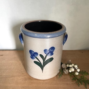 May include: A cylindrical stoneware crock featuring a speckled beige body, a blue rim, and two side handles. The front is decorated with a hand-painted blue floral motif with green leaves. The interior is dark, and the vessel has a smooth finish.