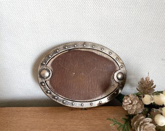 May include: Oval-shaped belt buckle with a brown leather center and a silver-colored metal frame. The frame has a series of small rivets around the edge and decorative rounded accents. The buckle is displayed on a wooden surface.
