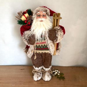 May include: A decorative Santa Claus figure, dressed in a red coat with faux fur trim and brown pants. He holds wooden skis and a sack of gifts. His sweater has a green and red pattern. The figure has a long white beard and is wearing glasses.