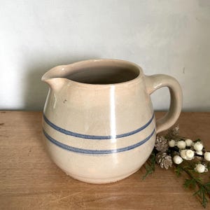 Puede incluir: Una jarra de cerámica color crema con dos rayas azules horizontales. La jarra tiene un cuerpo redondeado, un pico y un asa curva. Se coloca sobre una superficie de madera con elementos decorativos.