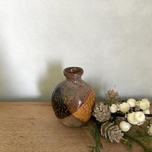 May include: A small, brown and black ceramic vase with a geometric pattern. The vase is sitting on a wooden surface.