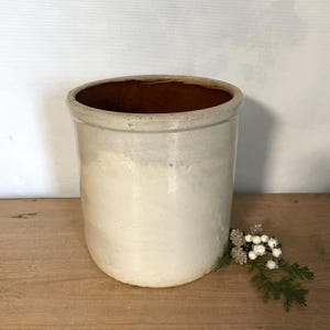 Może przedstawiać: Cylindryczny ceramiczny garnek o gładkiej, kremowobiałej szkliwionej powierzchni zewnętrznej i kontrastującym ciemnobrązowym wnętrzu. Naczynie posiada gruby, zaokrąglony brzeg oraz proste ścianki, co nadaje mu prosty i funkcjonalny wygląd.