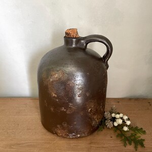 May include: A dark brown ceramic jug with a rounded body, a single side handle, and a cork stopper. The surface of the jug features a weathered, mottled texture with variations in color, showing signs of age and use across its exterior.