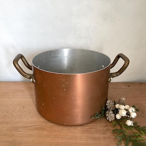 May include: A copper-colored cooking pot with two brass handles. The interior is silver-colored. The pot is sitting on a wooden surface. Decorative pinecones and greenery are placed near the pot.