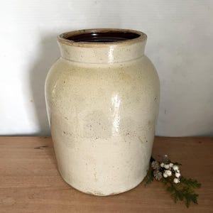 May include: A cream-coloured stoneware crock with a dark brown rim. The cylindrical jar has a wide opening and a slightly tapered body. The vintage kitchenware is displayed on a wooden surface. A classic piece.