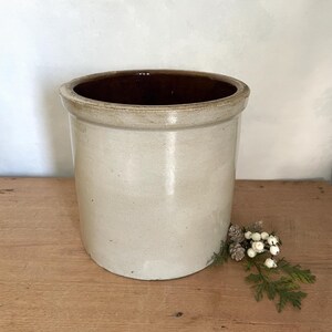 May include: A cylindrical stoneware crock featuring a smooth, light beige exterior glaze and a contrasting dark brown interior glaze. The vessel has a thick, reinforced rim and a flat base, presenting a simple, functional design for storage.