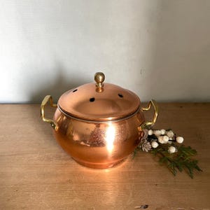 May include: A polished copper pot with a rounded body, two brass side handles, and a matching domed lid featuring a central brass knob and four small ventilation holes. The metallic surface reflects light, showcasing a warm, reddish-orange hue.