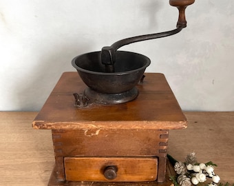 May include: An antique coffee grinder with a dark metal bowl and crank arm, mounted on a wooden base with a small drawer. The wood has a warm, aged tone, and the grinder shows signs of wear.