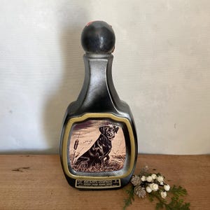 May include: A black ceramic decanter bottle with a rounded stopper, featuring a central rectangular label with a gold border. The label depicts a black dog in a grassy field with cattails. Text on the label reads BEAM'S CHOICE KENTUCKY STRAIGHT BOURBON WHISKEY.