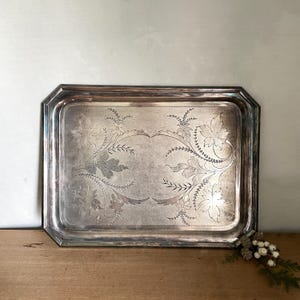 Könnte beinhalten: Ein rechteckiges silberfarbenes Metalltablett mit abgeschrägten Kanten und polierter Oberfläche. Die Mitte des Tabletts zeigt ein kompliziertes, geätztes Blumenmuster mit symmetrischen Blatt- und Rankenmotiven auf der Metalloberfläche.