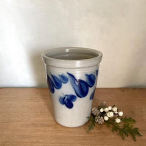 May include: A cylindrical stoneware crock featuring a speckled light grey base glaze and hand-painted cobalt blue teardrop or petal motifs encircling the upper portion of the vessel. The rim is smooth and slightly flared with a glossy finish.