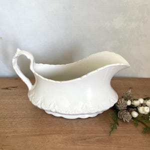 Puede incluir: Una salsera de porcelana blanca con asa curva y borde festoneado. La salsera tiene un patrón floral en relieve decorativo alrededor de la base. El asa es curva y tiene un diseño texturizado. La salsera está sobre una superficie de madera.