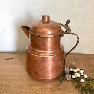 Puede incluir: Una tetera de cobre con tapa y asa curva. La tetera tiene un pico y un accesorio decorativo para el asa. El cobre tiene un acabado brillante. La tetera está sobre una superficie de madera.