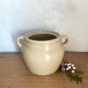 Peut inclure: Un pot en céramique de couleur crème avec une finition lisse et brillante, doté d'une large ouverture et de deux anses latérales arrondies. Le récipient présente une forme ventrue et se tient seul, arborant un vernis neutre et uni.