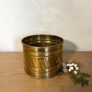 May include: A cylindrical brass planter featuring a polished metallic finish and a decorative pattern of diagonal grooves etched into the exterior surface. The container has a wide, open top and a smooth, flat base, presenting a reflective gold tone.