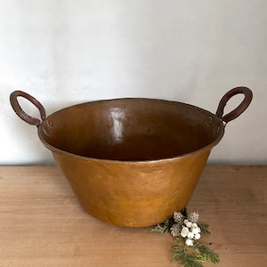 May include: A large, copper bowl with two rounded handles. The bowl is sitting on a wooden surface.
