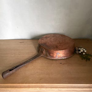 May include: A round copper pan with a long, dark metal handle attached by two rivets. The pan has a weathered, metallic finish with visible signs of use and oxidation. A small decorative sprig of white berries and green leaves sits nearby.