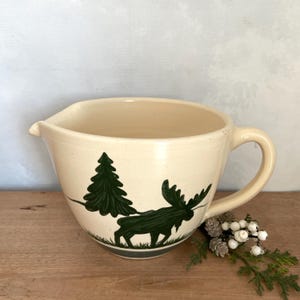 May include: Cream-colored ceramic mixing bowl with a spout and handle. The bowl features a green illustration of a moose and a pine tree. The bowl sits on a wooden surface.