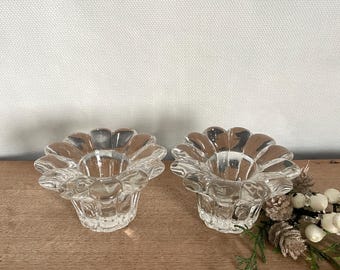 May include: Two clear glass candle holders shaped like flowers with multiple petals. The candle holders are sitting on a wooden surface.