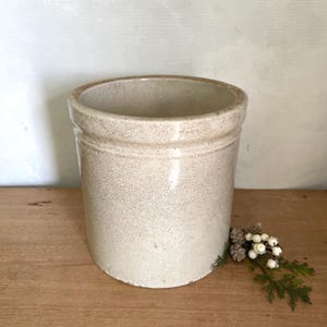 May include: A cylindrical ceramic crock featuring a light beige glaze with a visible crackle finish throughout the surface. The vessel has a thick, rounded rim and a straight-sided body, presenting a neutral, textured appearance for storage.