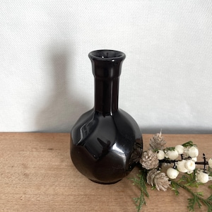 Puede incluir: Un jarrón de cerámica negro con cuerpo redondo y cuello estrecho. El jarrón tiene un patrón geométrico y un acabado brillante.