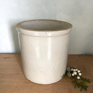 May include: A cylindrical off-white ceramic crock with a smooth, glazed finish and a thick, rounded rim. The vessel features a simple, unadorned exterior surface and a wide, open top, presenting a sturdy and functional design for storage or display.