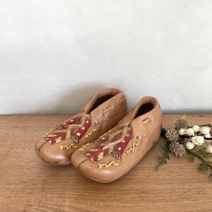 Puede incluir: Un par de mocasines de cerámica marrón con diseños geométricos rojos y amarillos. Los mocasines están sobre una superficie de madera.