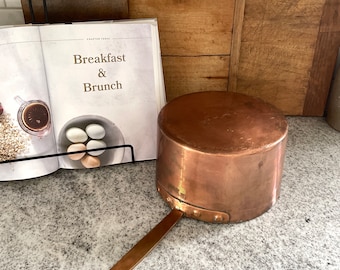 May include: A copper pot with a long handle and a rounded bottom. The pot is sitting on a gray countertop. The pot is shiny and has a rustic look.
