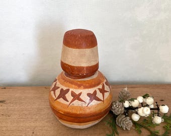 May include: A handcrafted, decorative ceramic bottle with a matching stopper. The bottle is painted with a warm color palette of brown, tan, and orange, featuring a starburst pattern. The stopper has a similar design. The bottle is displayed on a wooden surface.