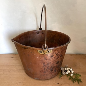May include: A weathered copper bucket with a tapered body, a rounded rim, and a sturdy metal swing handle attached by brass-coloured rivets. The exterior surface displays a rich, aged patina with dark spots and signs of oxidation throughout.