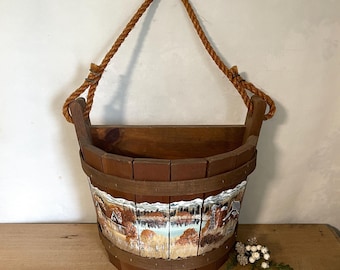 May include: A semi-circular wooden wall planter featuring a hand-painted landscape scene of trees, mountains, and a body of water. The planter is constructed from vertical brown wooden slats with a thick, natural-colored rope handle for hanging.