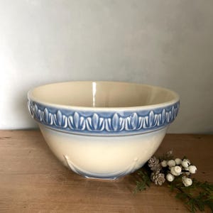 May include: A cream-colored ceramic mixing bowl featuring a wide, embossed blue band around the upper rim with a repeating leaf-like pattern. The bowl has a smooth, rounded base and a glossy finish, showcasing a classic design with subtle blue accents.