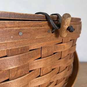May include: A brown woven basket with a wooden handle and a black leather strap. The basket is made of natural wood and has a rustic look.