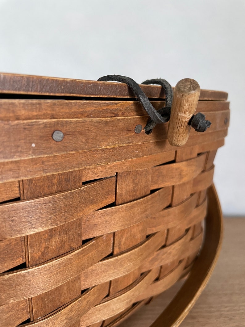 May include: A brown woven basket with a wooden handle and a black leather strap. The basket is made of natural wood and has a rustic look.
