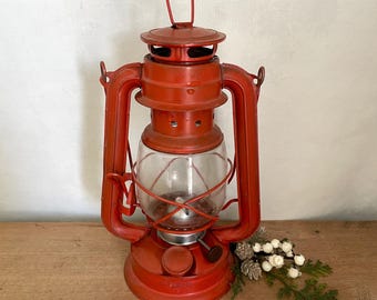 May include: A red metal hurricane lantern featuring a clear glass globe, a top carrying handle, and a side adjustment knob. The lantern has a circular base and a protective wire cage surrounding the central glass section for light output.