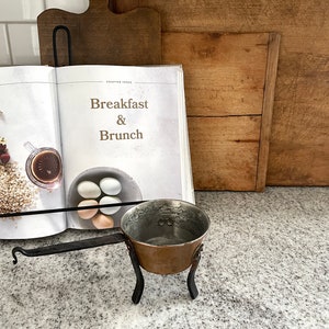 May include: A small copper pot with a black metal handle and stand. The pot is sitting on a gray countertop. The pot is in focus, and the background is blurred. The pot is a kitchen utensil.