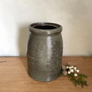 May include: A cylindrical stoneware crock featuring a dark gray, mottled glaze with a glossy finish. The vessel has a thick, rounded rim and a straight body. A small sprig of green cedar with white berries is positioned next to the base of the crock.