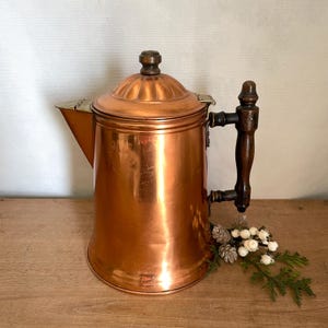 May include: A polished copper coffee pot featuring a tall, cylindrical body, a wide spout, and a domed lid with a dark brown wooden finial. The pot includes a matching dark brown wooden handle attached with brass hardware to the side.