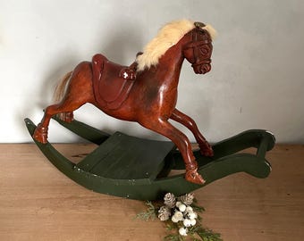 May include: A wooden rocking horse with a dark brown body, a saddle, and a cream-colored mane and tail. The horse is mounted on a green rocking base. Decorative pinecones and greenery are at the base of the horse.
