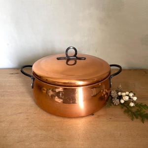 May include: A polished copper pot with a matching lid, featuring two curved black side handles and a circular black handle on top. The pot has a smooth, reflective surface and a rounded shape, showcasing a warm metallic finish throughout.