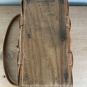 May include: A Longaberger basket with a wooden lid and a leather handle. The lid has a label that reads "Longaberger Baskets Handwoven U.S.A." and "1486".
