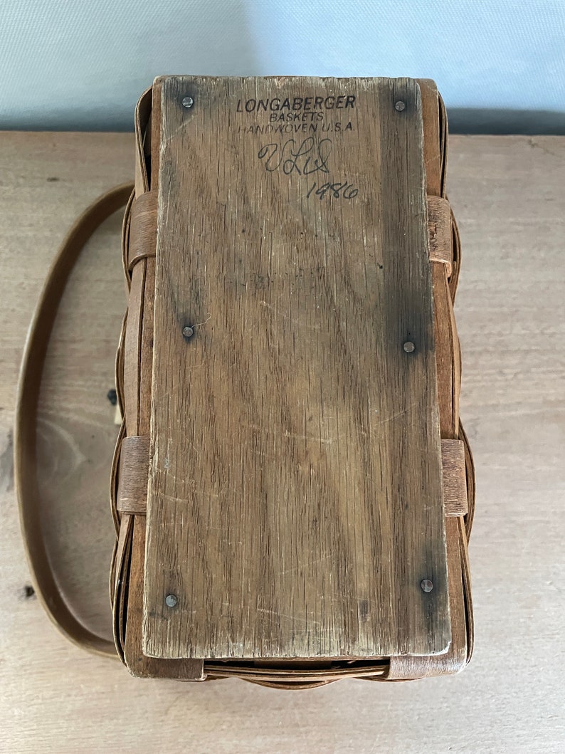 May include: A Longaberger basket with a wooden lid and a leather handle. The lid has a label that reads "Longaberger Baskets Handwoven U.S.A." and "1486".