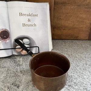 May include: A small copper saucepan with a wooden handle. The saucepan is empty and has a shiny, slightly tarnished surface.