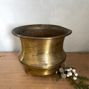 Może przedstawiać: Antyczna mosiężna misa z szerokim brzegiem i zaokrąglonym korpusem. Misa ma złoty odcień ze śladami wieku i zużycia. Stoi na trzech małych nóżkach i jest eksponowana na drewnianej powierzchni.