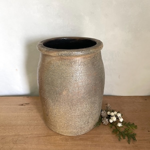 May include: A cylindrical stoneware crock featuring a textured, mottled brown and tan exterior finish with a dark interior. The vessel has a thick, rounded rim and a straight body that tapers slightly toward the base, showcasing a rustic appearance.